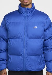 Mėlyna pūkinė striukė su aukštu apykakle, užtrauktuku ir Nike logotipu. Pagaminta iš siuvinėto audinio su sklandžia tekstūra ir pūsta forma.