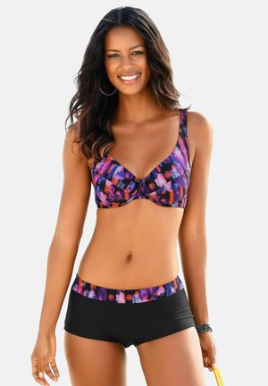 Lächelnde Frau mit langen Haaren, die ein bunt gemustertes Bikini-Oberteil und schwarze Badeshorts mit passendem Bund trägt, hält ein gelbes Objekt in der Hand.