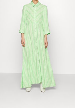 Persoon draagt een lange, lichtgroene jurk met patroon, 3/4-mouwen en kraag, gecombineerd met witte laarzen met puntige neus, staand tegen een effen achtergrond.