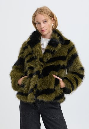 Femme aux cheveux blonds portant une veste en fausse fourrure rayée verte et noire par-dessus une chemise blanche et un jean noir, les mains dans les poches de la veste.