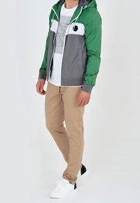 Zielono-szara kurtka zapinana na zamek z kapturem, z białym paskiem na wysokości klatki piersiowej. W połączeniu z beżowymi spodniami i białymi trampkami.