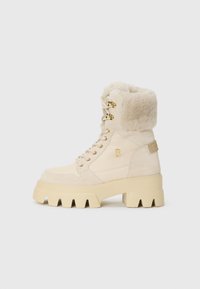 Beige enkelboot met een fluffy manchet, voorzien van gouden veterogen en veters, en een chunky platformzool, tegen een effen witte achtergrond.