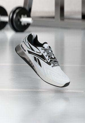 Witte en zwarte Reebok hardloopschoen met patroonveters die boven een sportschoolvloer zweeft, met een wazige dumbbell op de achtergrond.