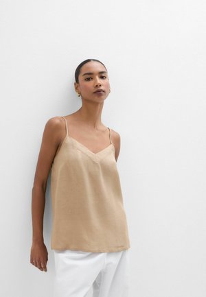 Kobieta z włosami zaczesanymi do tyłu, nosząca złote kolczyki koła, beżowy top bez rękawów z cienkimi ramiączkami i białe spodnie, stojąca przy białej ścianie.