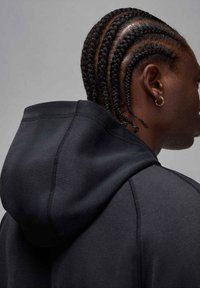 Czarna bluza z kapturem o fakturowanej powierzchni, z kapturem na sznurki. Włosy ułożone w wiele ciasnych warkoczy z tyłu, widoczna złota kolczyk w kształcie koła.