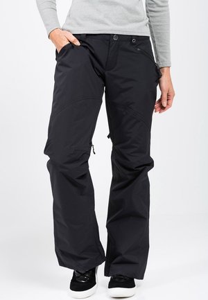 Pantalons noirs imperméables à coupe large, avec poches zippées, genoux renforcés et texture lisse. Portés avec des baskets noires.