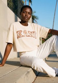 Lühikeste juustega noor inimene, kes kannab beeži lühikest t-särki ja valgeid pükse, istub päikesepaistes kõnniteel palmipuude lähedal.