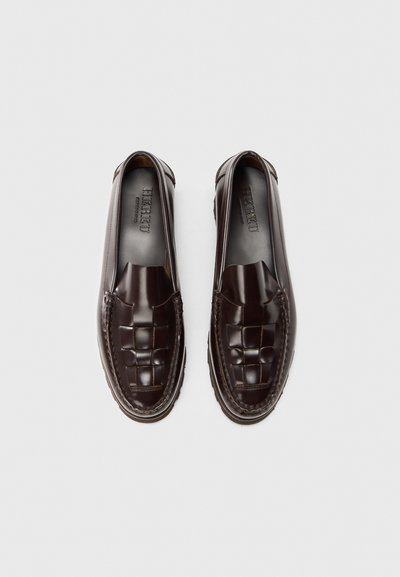 Pereche de mocasini din piele maro închis, cu model împletit în partea de sus, talpă plată și branț cu brand, așezați unul lângă altul pe un fundal alb.