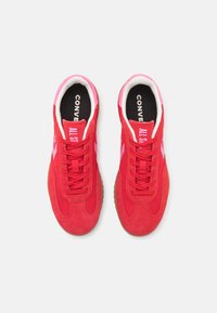 Røde sneakers med en øvre del i ruskind og tekstil, prydet med lyserøde detaljer, runde snørebånd og en beige gummisål. "CONVERSE ALL STAR" logoet er synligt.