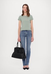 Camiseta ajustada de color verde grisáceo con texto, vaqueros azules, bolso de cuero negro con detalle de lazo, zapatos negros de punta, fondo minimalista.