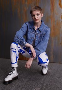 Chaqueta de mezclilla en azul claro, pantalones blancos con patrones abstractos en azul y botas blancas con cordones y suela negra gruesa. Fondo texturizado.
