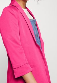 Blazer fucsia con vestibilità sartoriale, rever a punta e maniche arrotolate, indossato sopra una camicia di denim con bottoni. Texture morbida, design minimalista.