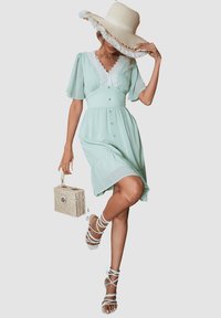Vestido verde claro con escote en V, detalles de encaje, mangas abullonadas y botones al frente. Combinado con un sombrero de paja de ala ancha y sandalias blancas.
