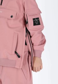 Roze, met rits sluitende jas gemaakt van gladde stof heeft een zak op de borst, geribbelde manchet en labelaccenten. Close-up van de arm- en zakdetails.