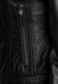 Uski crna kožna jakna u krupnom planu prikazuje bočni patentni zatvarač, vidljive šavove i različite teksture na materijalu. Zatvarač ima metalni povlačnik.