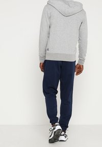 Sweatshirt à capuche gris avec des poignets côtelés, associé à un jogging bleu marine. Chaussures de sport noires et blanches. Logo visible sur le sweatshirt.