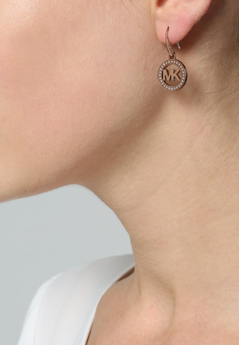 Des boucles d'oreilles créoles en or rose présentent un pendentif circulaire avec un logo "MK" gravé, entouré d'un anneau de cristaux transparents pour une touche de détail ajoutée.