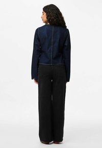 Giacca di jeans blu scuro con un design aderente, maniche lunghe e cuciture in vista, abbinata a pantaloni neri a gamba ampia.