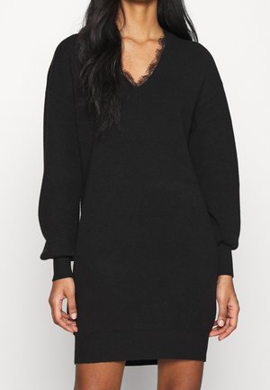 Woman wearing a black long-sleeve knit dress with a V-neckline featuring delicate lace trim, standing against a plain background.