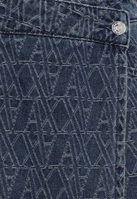 Tissu en denim foncé avec un motif géométrique de formes imbriquées et des coutures visibles. Comprend un accent en bouton argenté sur la ceinture.