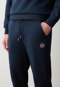 Pantaloni della tuta blu navy realizzati in tessuto morbido, dotati di una vita regolabile con hardware argentato e una patch rotonda con logo sulla coscia sinistra.