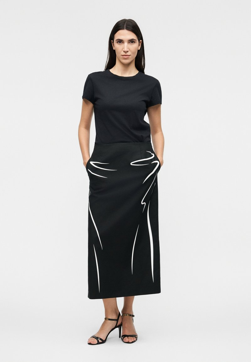 Woman with long dark hair stands wearing a black short-sleeve top and a black midi skirt with white abstract line patterns, hands in pockets.