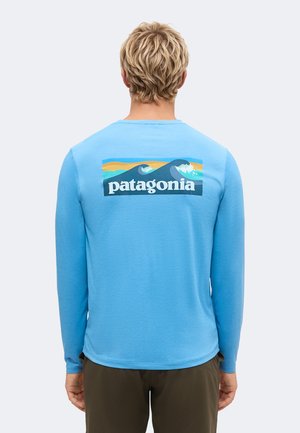 Persona rubia de espaldas que lleva una camisa azul de manga larga con un gráfico de onda colorida y el texto "patagonia" en la parte trasera.