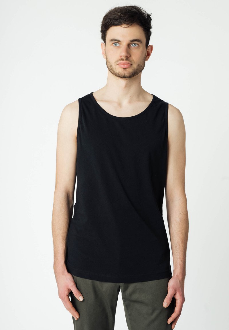 Schwarz ärmelloses Baumwoll-Tanktop mit rundem Halsausschnitt, das eine glatte Textur und eine lockere Passform aufweist, kombiniert mit olivgrünen Hosen.