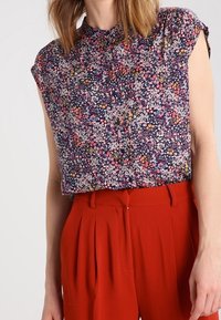 Blusa floral de manga corta, fondo azul marino, pequeñas flores rosas y amarillas, detalle de plisado en el escote, combinada con pantalones rojos de cintura alta.