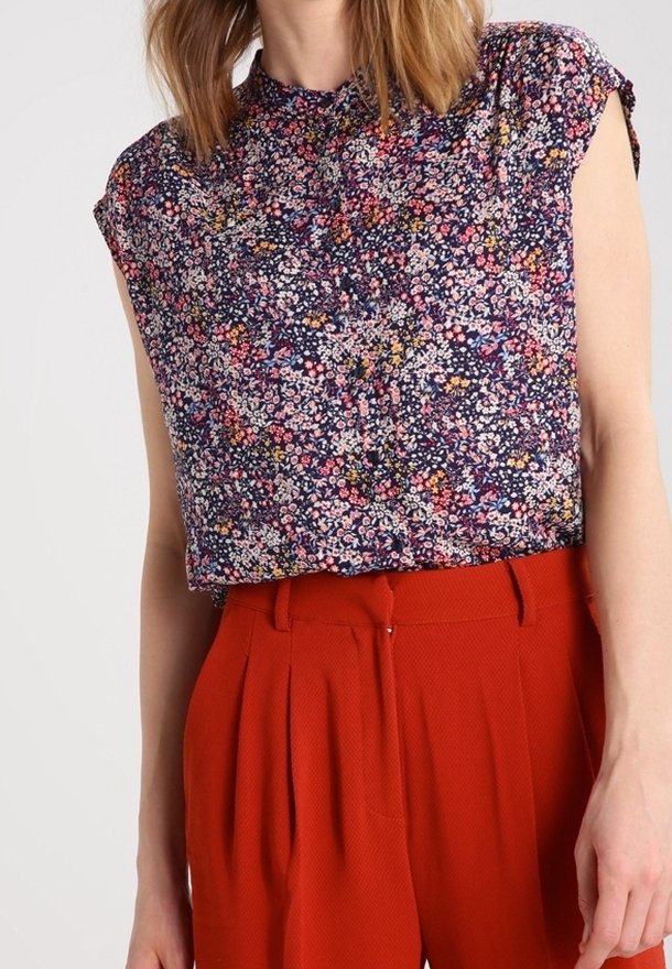 Blusa floral de manga corta, fondo azul marino, pequeñas flores rosas y amarillas, detalle de plisado en el escote, combinada con pantalones rojos de cintura alta.