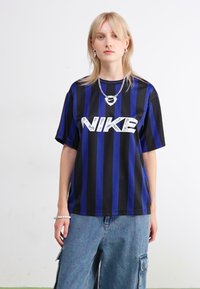 Camiseta Nike a rayas negras y azules con un ajuste holgado, que presenta un gran logo plateado en la parte frontal. El modelo lleva pantalones vaqueros casuales.