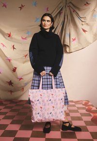 Moteris juodu golfu, mėlynu languotu sijonu ir juodais batais laiko didelę rožinę nešiojamą krepšį su spalvotais šunų raštais ant rožinės languotos grindų dangos.