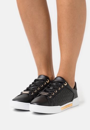 Jambes portant des baskets en cuir noir avec des œillets dorés, des semelles blanches et des accents de marque, posant sur un fond blanc.