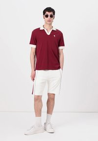 Burgundi polo marškiniai su balta apykakle ir rankogaliais, derinami su baltais šortais su šoniniais juostelėmis. Modelis dėvi saulės akinius ir baltus sportbačius.