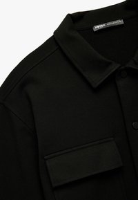 Camisa negra con cuello, cierre de botones y un gran bolsillo en el pecho. La tela parece texturizada y está hecha de un material suave.