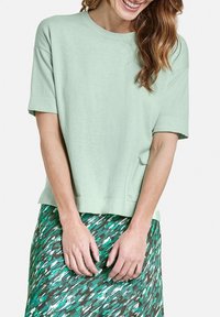 T-shirt verde chiaro a maniche corte con scollatura rotonda; tessuto morbido e testurizzato. Abbinato a una gonna multicolore e a fantasia in tonalità di verde e marrone.