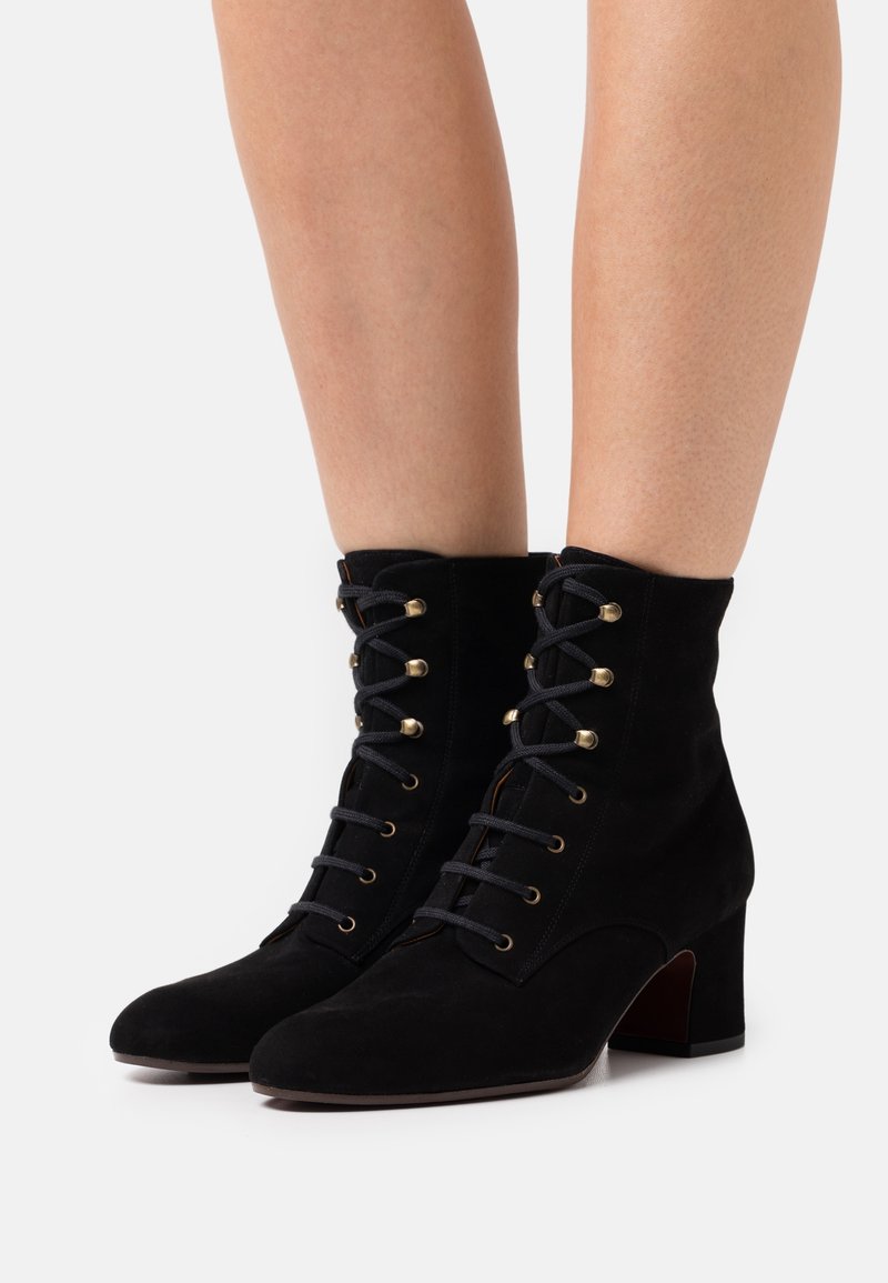 Stivaletti neri in suede con tacco alto, caratterizzati da occhielli e lacci dorati, design strutturato e punta rotonda.