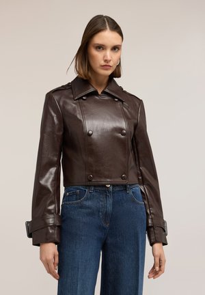 Donna con giacca doppiopetto in pelle marrone scuro corta con polsini con fibbia e jeans blu a vita alta su sfondo semplice.