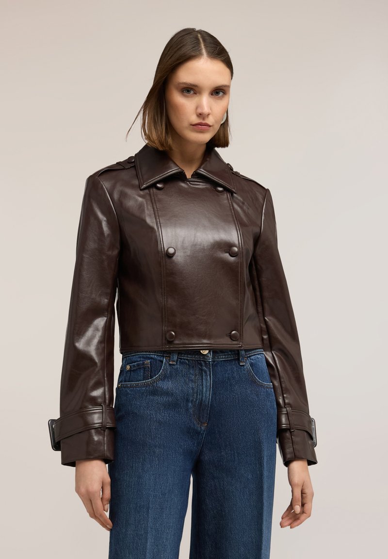 Femeie purtând o jachetă scurtă din piele maro închis, cu două rânduri de nasturi și manșete cu cataramă, și blugi albaștri cu talie înaltă, pe un fundal simplu.