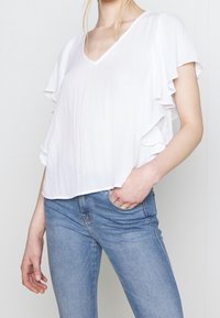 Top bianco a maniche corte con scollatura a V, caratterizzato da dettagli arricciati lungo le maniche. Abbinato a jeans slim-fit blu chiaro. Tessuto morbido.