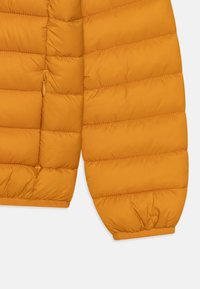 Højre ærme og side af en orange quiltet pufferjakke med synlig lynlås lomme og elastisk manchet på hvid baggrund.