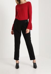 Femme portant un haut rouge à manches longues avec des poignets évasés, un pantalon noir taille haute avec une boucle sur le côté, et des escarpins noirs à bout pointu, debout.