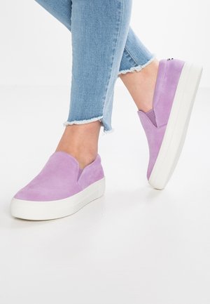 Person iført lyseblå jeans med frynset kant og lavendel slip-on platformsneakers på et hvidt gulv.
