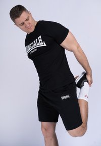 Zwarte Lonsdale T-shirt en shorts, met wit logo; model strekt been, draagt zwarte schoenen en witte sokken met rode accenten.