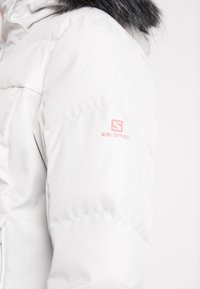 Manche de veste d’hiver matelassée blanche avec logo Salomon rose et capuche en fausse fourrure noire visible sur le bord supérieur.