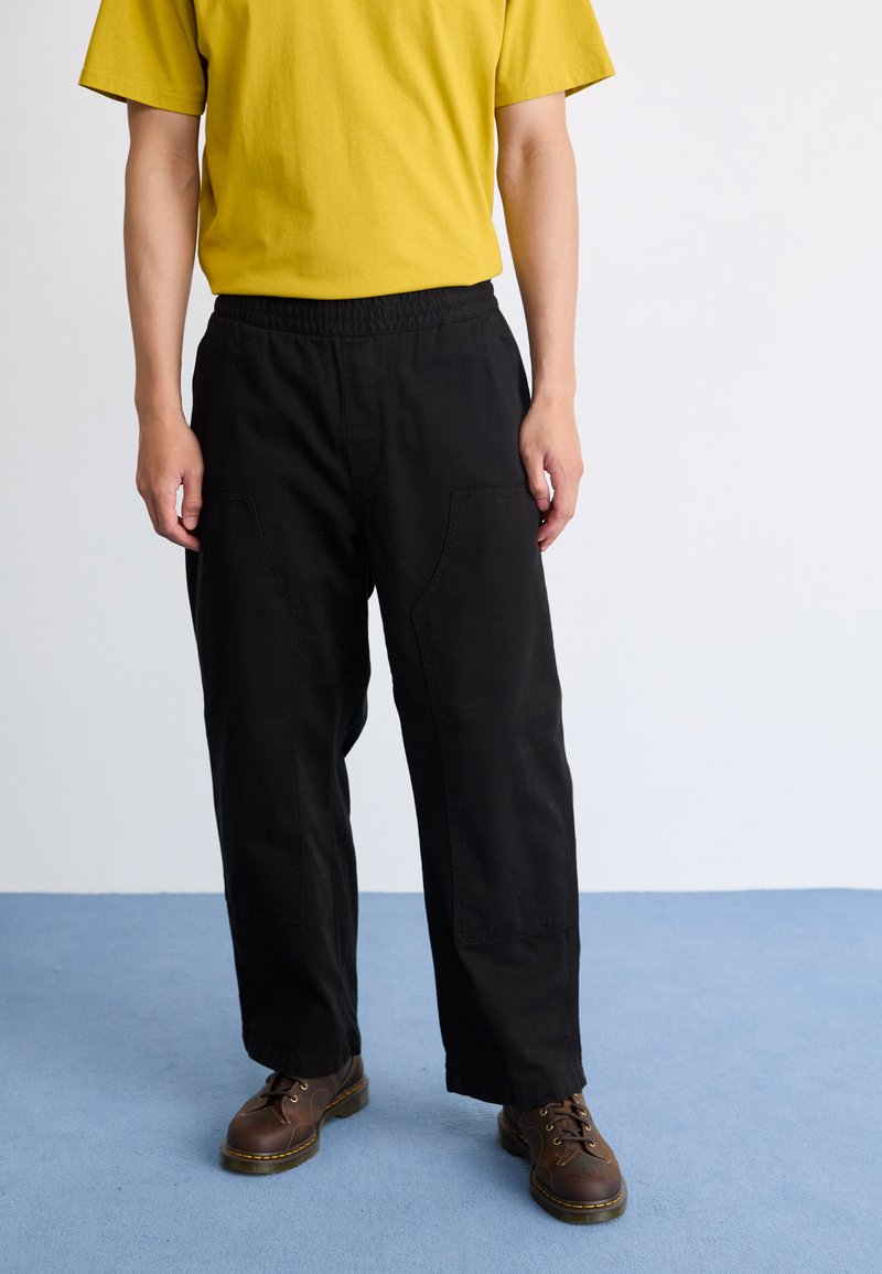 Pantalones negros de cintura elástica con un corte relajado, con bolsillos laterales. Combinados con una camiseta de algodón amarilla y zapatos marrones de cordones.
