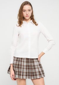 Άσπρο πουκάμισο με κουμπιά και μακριά μανίκια, συνδυασμένο με καρό πλισέ φούστα καφέ και λευκού χρώματος, που παρουσιάζει λεία υφή και κλασικό σχεδιασμό.