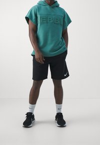 Turkos huva sweatshirt med NEBBI-logotyp, i kombination med svarta shorts. Modellen bär vita strumpor och svarta sneakers med grå detaljer.