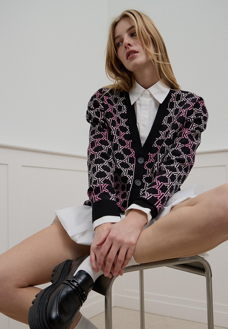 Schwarze und rosa gemusterte Strickjacke mit Puffärmeln, Knopfverschluss und gerippten Bündchen, getragen über einem weißen Hemd und kombiniert mit schwarzen Schuhen.