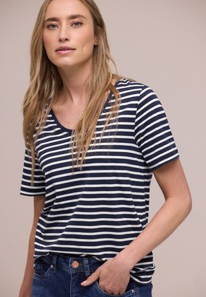 Mujer con cabello largo y rubio, vistiendo una camiseta de manga corta con rayas azul marino y blancas y jeans azules, con la mano en el bolsillo, fondo neutro.